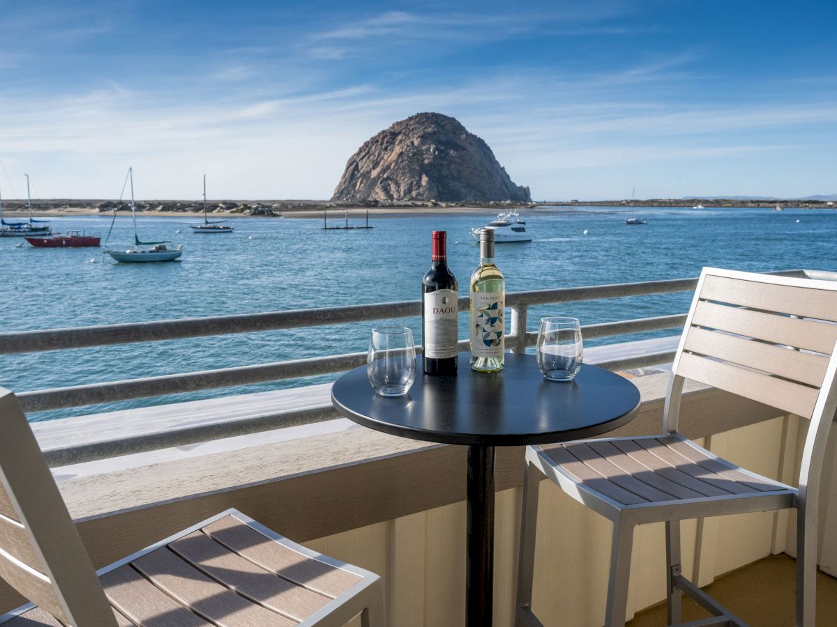A serene coastal view featuring a table with two wine bottles, empty glasses, and chairs overlooking boats on the ocean and a large rock formation.