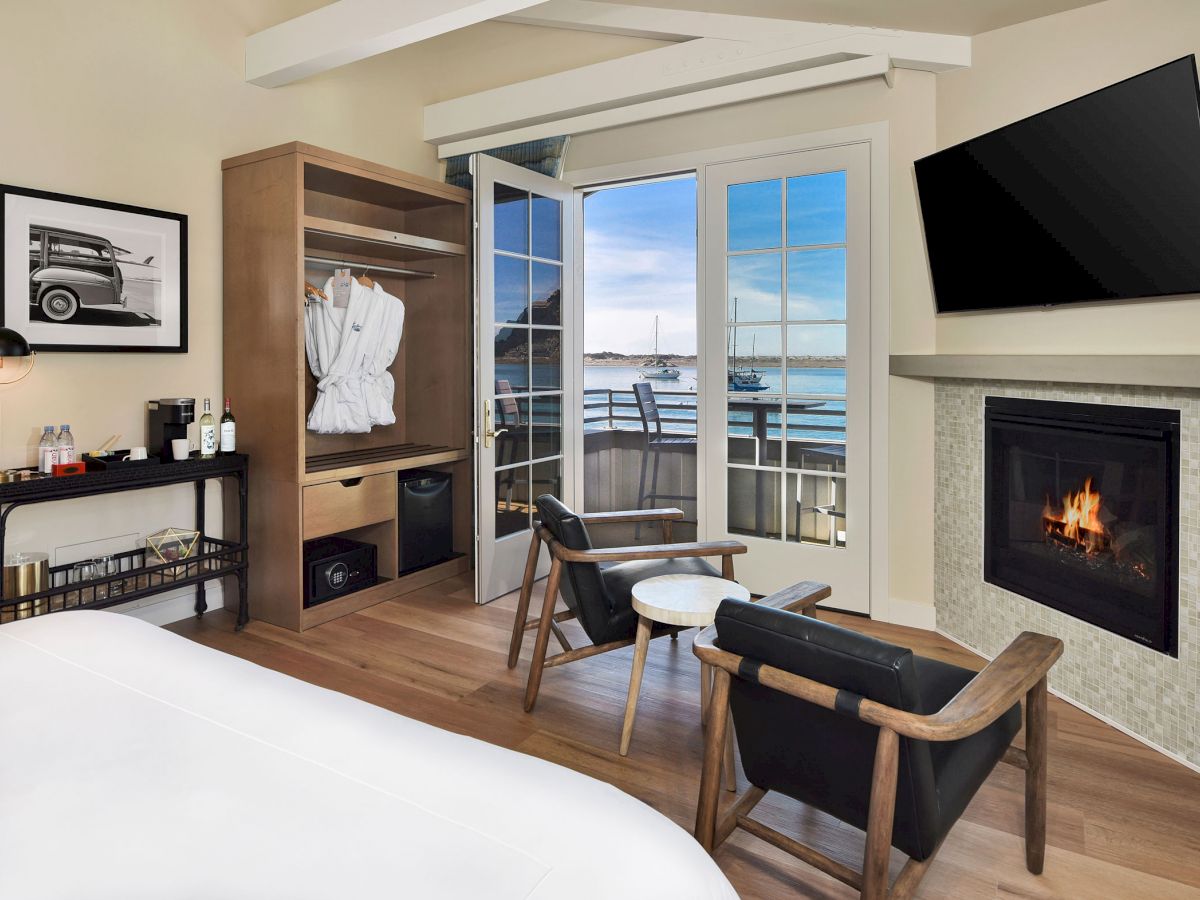A cozy room with a bed, open closet with robes, two chairs, a fireplace, TV, and a view of a marina through French doors completes the setup.